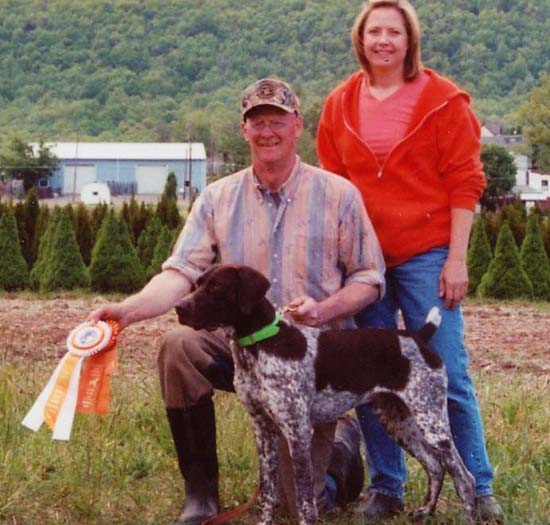 A Fogelhund German Shorthaired Pointer stands with its proud owners after winning two ribbons.