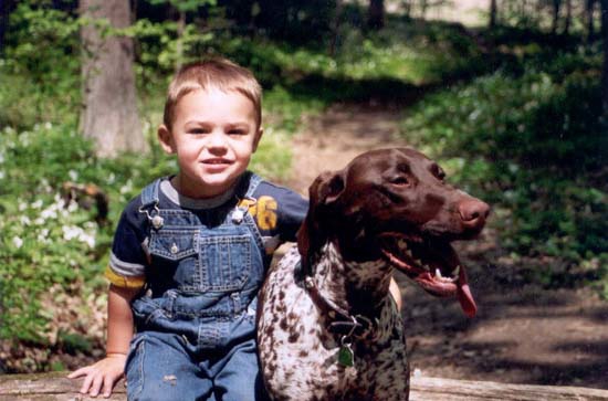 A little boy in blue jean overalls with his arm around a Fogelhund German Shorthaired Pointer, with its tongue hanging out.